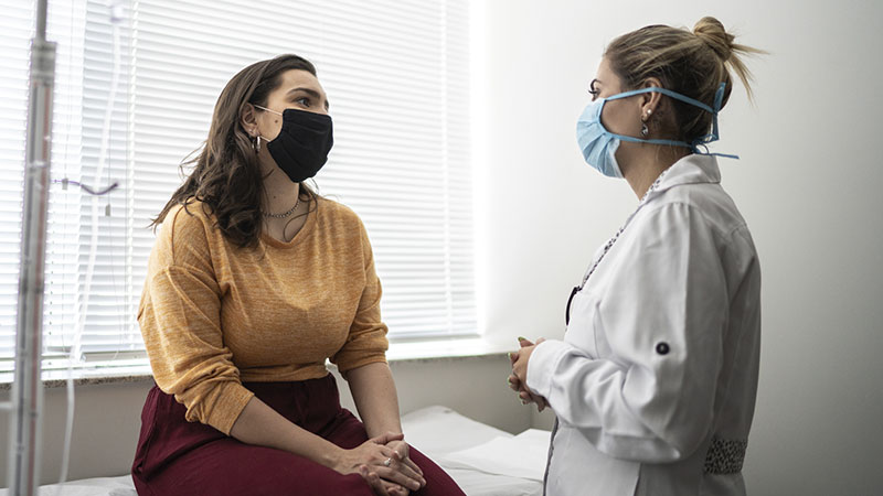 Médica conversando com paciente mulher que está sentada e de máscara